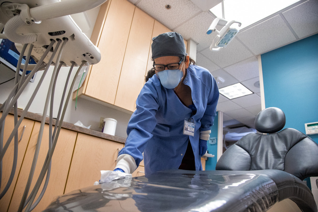 A female dental assistant wiping down the medical chair after spraying it with surface disinfectants and cleaners in a dental setting.